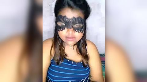 Media: Video of a young woman with light brown skin, wearing a black lace masquerade mask, blue striped tank top, and brown hair, indoors with blurred background.