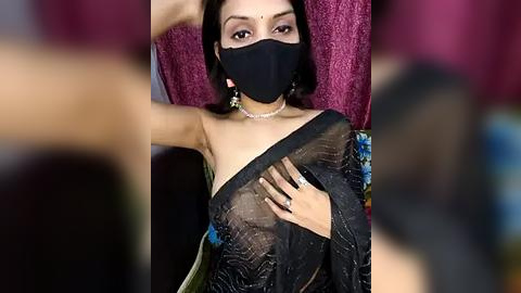 Media: Video of a woman in a black saree with a sheer, blue floral pattern, wearing a black face mask, sitting indoors with a red velvet backdrop.