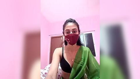 Media: Video of a South Asian woman with medium brown skin and black hair, wearing a black bra and a green saree with a red face mask, standing in a pink room with a door and window.