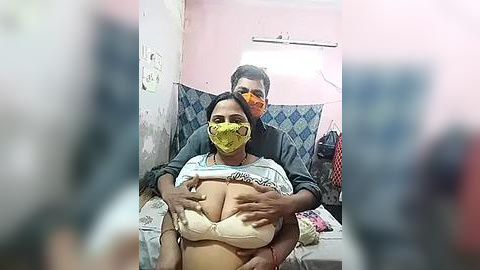 Media: Video of a man and woman in a hospital room. The woman, wearing a yellow mask and a white bra, is seated. The man, wearing glasses and a mask, stands behind her, holding her breasts.