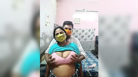 Media: Video of a young woman with dark hair, wearing a yellow face mask and a pink bra, being lifted by a man in a white shirt in a pink-painted room with a blue-patterned bedspread.