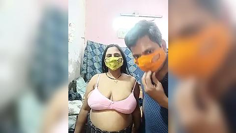 Media: Video of a woman with medium-dark skin, wearing a yellow face mask, pink bra, and patterned skirt, standing next to a man with dark skin, wearing an orange face mask, in a small, dimly-lit room with peeling pink walls and a blue-patterned curtain.