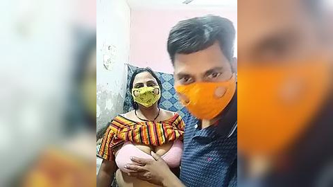 Media: Video of an Indian woman with a yellow mask, wearing a colorful off-shoulder top, covering her breasts with a pink bra. A man with an orange mask stands beside her. Background shows a simple room with pink walls.