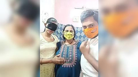 Media: Video of three individuals, two women in beige and blue traditional attire, and a man in a white shirt, wearing face masks, standing in a pink-walled room.