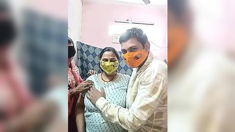 Media: Video of a man in an orange mask and beige shirt giving a vaccine to a woman in a light blue hospital gown with a yellow mask, in a pink-walled room with a blue patterned curtain.