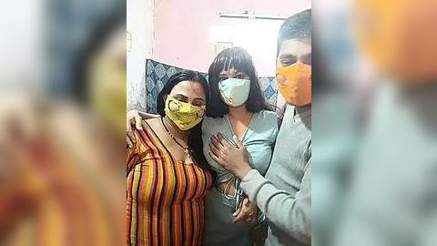 Media: Video of three women, wearing yellow face masks, standing in a dimly lit room with pink walls and blue-patterned tiles. One woman in a striped dress hugs another in a light blue top and a third in a grey sweater.