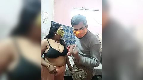 Media: Video of a man in a grey sweater and woman in a black bra and pink pants, both wearing face masks, standing in a dimly lit, cluttered room with peeling walls.