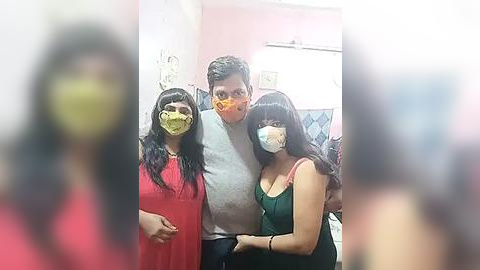 Media: Video of three people wearing yellow surgical masks; two women in red and green dresses, and a man in a gray shirt. Background shows a pink-painted wall and a white tiled floor.