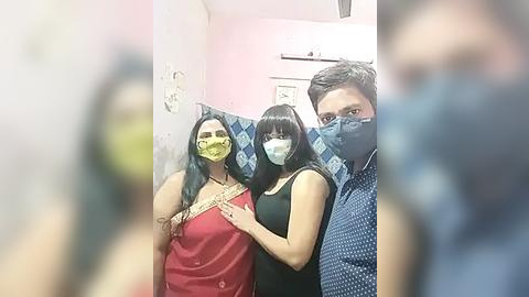 Media: Video of three people wearing yellow masks in a pink-walled room, with a man in a blue shirt partially visible in the background.