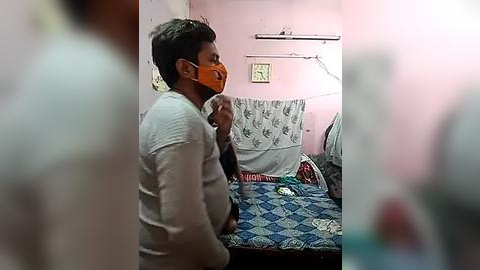 Media: Video of a man wearing a mask, sitting on a bed in a modest room with pink walls and a blue-patterned bedspread, holding a phone.