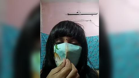 Media: Video of a person with long black hair and a light blue surgical mask, standing indoors with blue wallpaper and white ceiling. The image is blurry with a focus on the person\u2019s face.