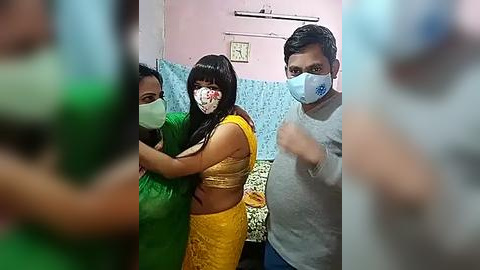 Media: Video of three people wearing face masks, standing in a room with pink and blue walls, one woman in a yellow saree and another in green, and a man in a grey shirt.