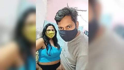 Media: Video of a man and woman wearing face masks in a dimly lit room with pink walls. The man has short dark hair, wearing a gray shirt, and the woman has long black hair, wearing a yellow mask and a blue crop top.