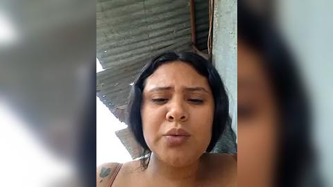 Media: A video of a young woman with medium brown skin and straight black hair, wearing a tank top, standing against a corrugated metal wall in a shanty town setting.