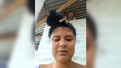 Media: Video of a young woman with medium brown skin, dark hair in a messy bun, wearing a light-colored top. Background shows a rustic, corrugated metal roof and white walls. She has a contemplative expression.