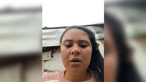 Media: A close-up video of a young woman with dark skin, medium build, and medium-length black hair, wearing a pink sweater, standing against a dilapidated, gray metal fence in an urban setting.