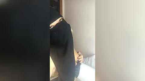 Media: Video of a person standing in a corner, partially obscured by a large, dark, rectangular object. The person has light skin and is wearing a black shirt. The background is a plain white wall.