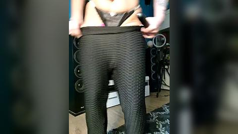 Media: Video of a woman pulling up her tight, textured black leggings, revealing her smooth, light-toned thighs. She has a slim build and is standing in a cluttered room with audio equipment in the background.