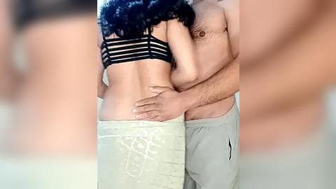 Media: Video of a dark-skinned woman with curly hair in a black bra and white pants, being held from behind by a light-skinned man in grey sweatpants, in a softly lit room.