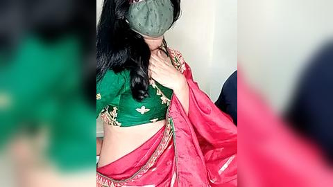 Media: Video of a woman with medium skin tone, wearing a green blouse with gold embroidery, a pink sari, and a green face mask, standing indoors with a blurred background.