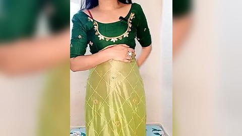 Media: Video of a woman in a green blouse with gold embroidery and a green dupatta, paired with a yellow salwar kameez featuring intricate gold embroidery. She stands indoors against a plain white wall, wearing a large ring.