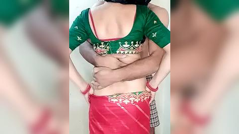 Media: Video of a woman in a green blouse with red trim, holding a man's bare back. She wears a red sari with a checkered border, standing against a blurred background.