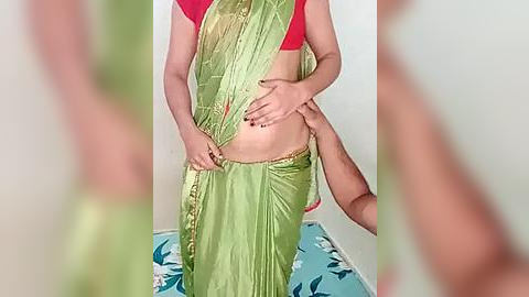 Media: Video of a person wearing a green and gold sari, red blouse, and white flowers, with another person's hands on their stomach. Background features a light blue tablecloth with white flowers.