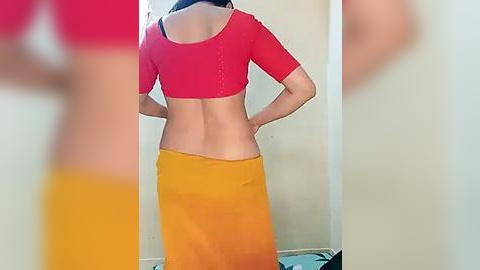 Media: Video of a woman with light skin, wearing a red crop top and a yellow skirt, standing with her back to the camera in a room with beige walls.