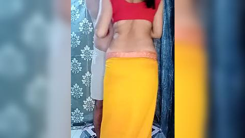 Media: A video showing a woman with light skin wearing a red crop top and bright yellow skirt, posing in a bathroom with white and blue patterned wallpaper and a tiled floor.