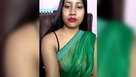 Media: A video of a young South Asian woman with long black hair, wearing a green saree, seated on a brown chair against a pastel wall. The image is slightly blurred.