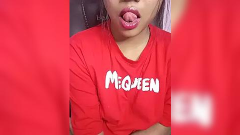 Media: A close-up video of a woman with medium brown skin, wearing a red t-shirt with the word \"MAGUEN\" in white, sticking out her tongue. The background is blurred.