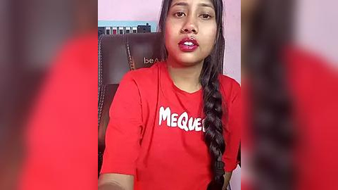 Media: Video of a young woman with long black hair, wearing a red T-shirt with \"MEGUEL\" printed in white, sitting in a chair. Background shows a blurred person with red hair.