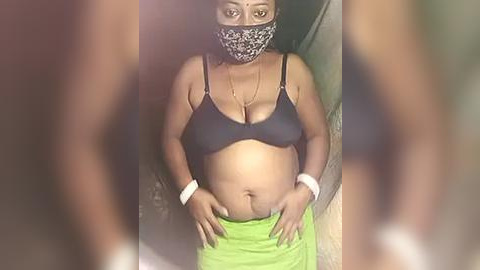 Media: Video of a pregnant Black woman in a black bra, leopard-print mask, and green skirt, with visible belly and wrists adorned with white bracelets, standing indoors.