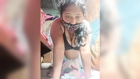 Media: Video of a South Asian woman with dark skin, wearing a black bra, patterned face mask, and sheer skirt, kneeling on a colorful rug in a cluttered room.