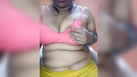 Media: Video of a plus-sized Black woman with a large chest, wearing a pink bra and yellow pants, standing in a dimly lit room with blurry background.