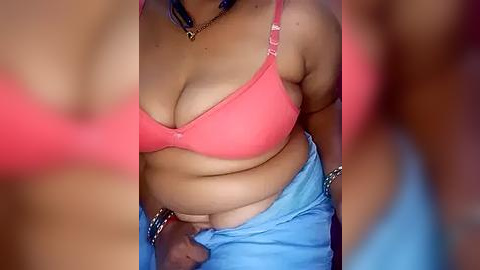 Media: Video of a plus-sized woman with light brown skin, wearing a pink bra, blue jeans, and a black necklace. The image is slightly blurred, with a focus on her midsection.