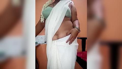 Media: Video of a dark-skinned woman wearing a white sari, a green bra, and a white necklace, standing in a room with orange walls and a bed visible in the background.