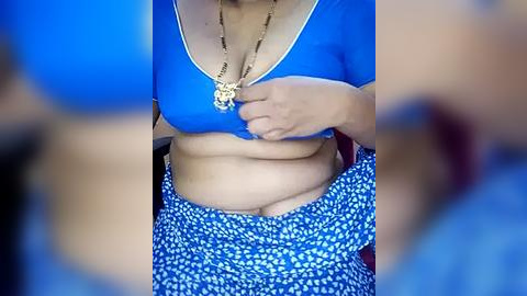 Media: Video of a woman with medium skin tone wearing a blue crop top and blue floral skirt, lifting her top to reveal her midriff and navel.