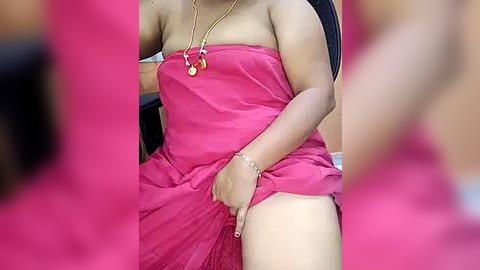 Media: A video of a woman with medium skin tone in a strapless, pink dress lifting the hem to reveal her thighs. She wears a gold necklace with a pendant and a bracelet. The background is blurred, focusing attention on her attire and jewelry.