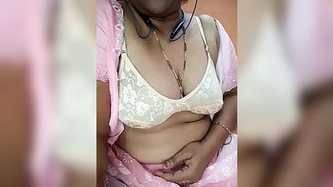 Media: Video of a light-skinned woman with medium breasts, wearing a pale pink saree, a white lace bra, and a gold chain necklace. She holds her stomach, wearing headphones. Background blurred.