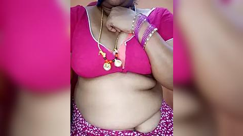 Media: A video of a plus-size woman with medium brown skin, wearing a bright pink crop top that accentuates her large breasts, and a patterned skirt. She accessorizes with gold necklaces and bracelets.