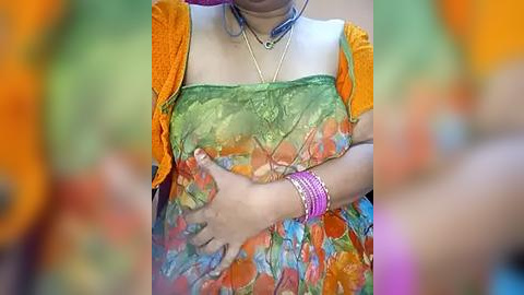 Media: A video of a woman with light skin tone, wearing an orange shawl over a colorful, floral-patterned blouse, accessorized with a gold necklace, bracelets, and a blue dog tag.