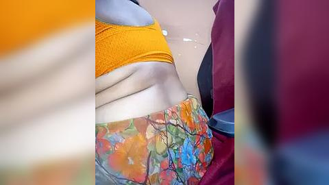 Media: Video of a woman's midsection, wearing a vibrant orange bra and colorful, patterned shorts, with blurred, colorful background.