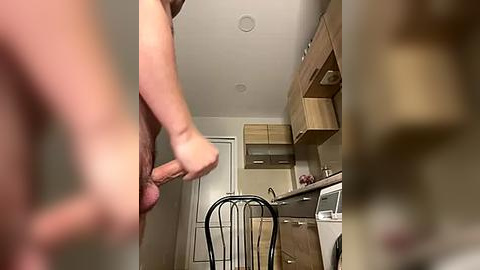 Media: Video of a nude man with a flaccid penis, standing in a modern kitchen with light wood cabinets, a black chair, and a microwave.