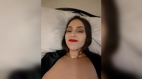 Media: A video of a fair-skinned woman with long dark hair, closed eyes, and red lipstick, lying on a white pillow in a black robe, with a blurred figure in the foreground.