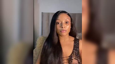 Media: Video of a young Black woman with long, straight black hair, wearing a black sequined top, standing in front of a gray headboard, blurred background, indoor setting.