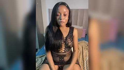 Media: Video of a young Black woman with long, straight black hair, wearing a black, lace-up mesh bodysuit, sitting on a bed in a dimly lit bedroom.