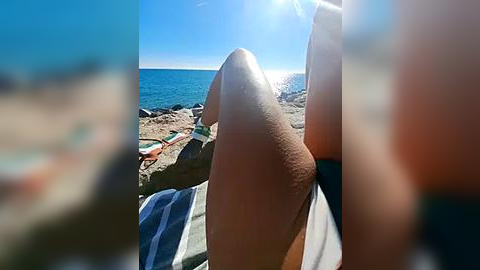 Media: A video captures a tan, muscular leg of a person reclining on a striped beach towel, with a clear blue sky and ocean backdrop.