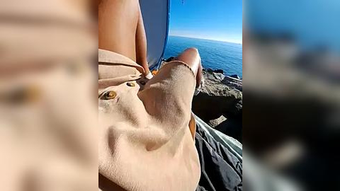 Media: Video of a shirtless person with fair skin, wearing a beige hat, lying on a rocky cliff overlooking a vast blue ocean. The sky is clear, with a slight blur in the foreground.