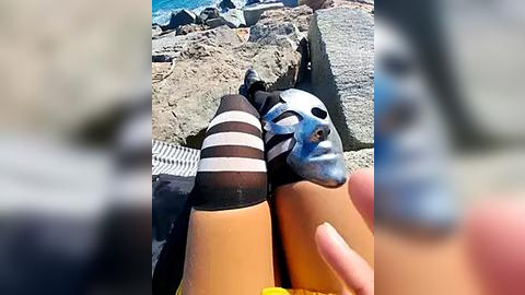 Media: Video of a person's hands holding two painted, black-and-white-striped, orange ice cream cones. Background shows rocky coastal terrain with blue sky and water. The cones have a surreal, cartoonish appearance.